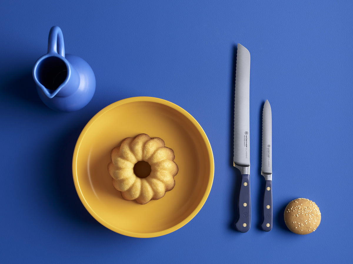 Classic 9" Precision Double-Serrated Bread Knife with a deep blue ergonomic handle and brass rivets, displayed next to a matching paring knife, a yellow plate with a bundt cake, and a blue pitcher on a blue background.