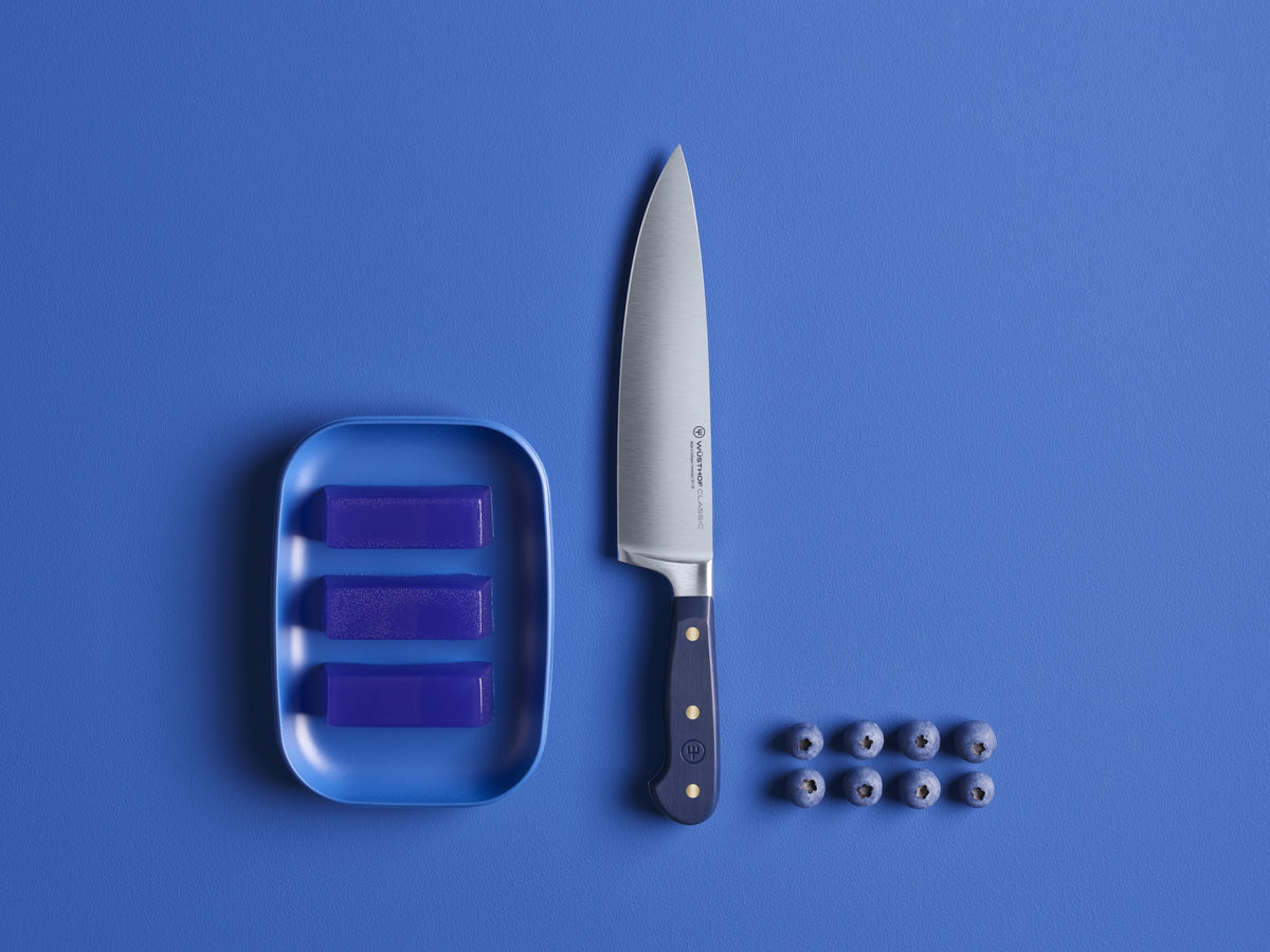 Classic 8" Chef's Knife with a polished forged German steel blade and a dark blue handle secured by three brass rivets, displayed beside a blue soap dish and fresh blueberries on a blue background.