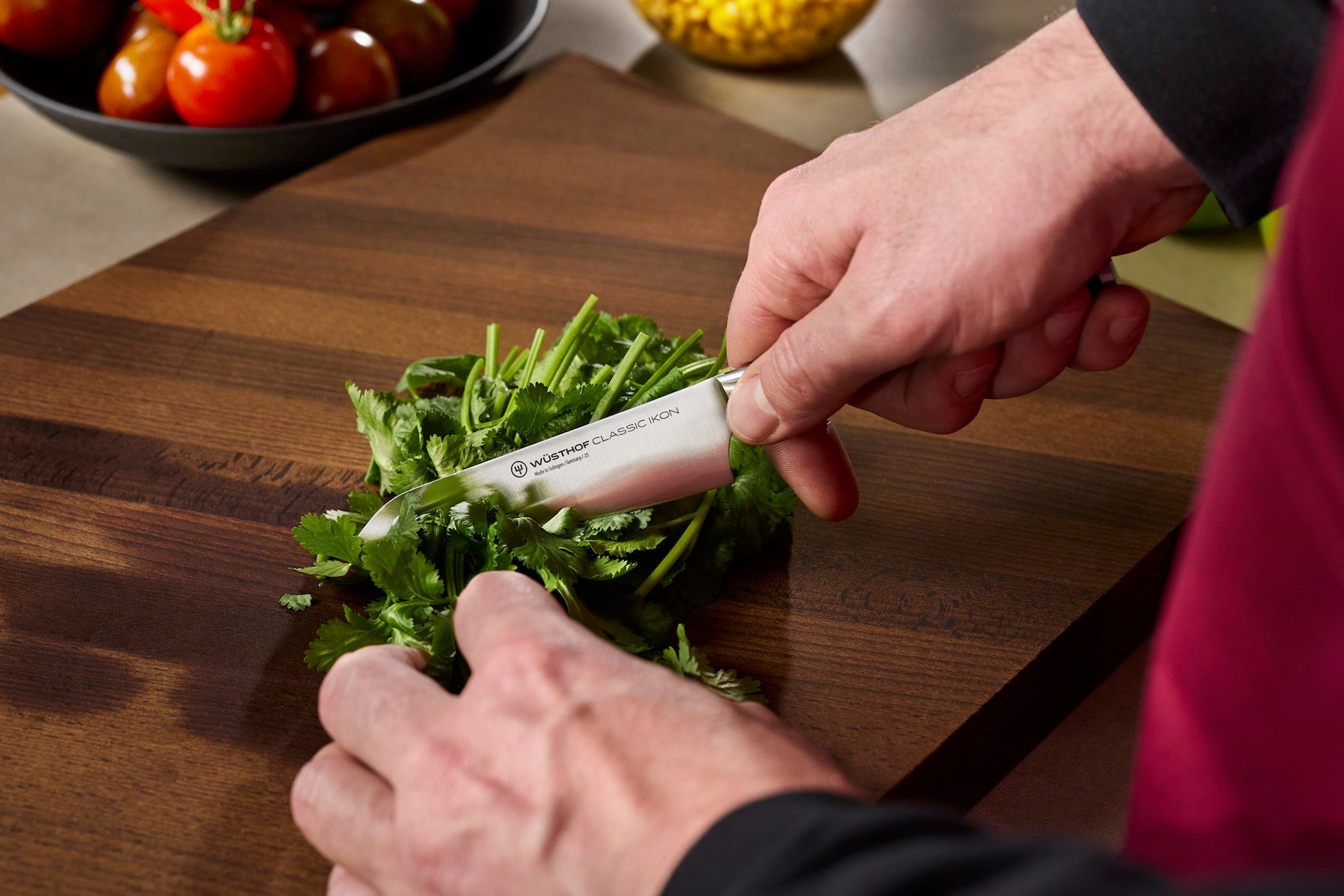A close-up of a person's hands using a Victorinox kitchen knife to chop green herbs on a dark wooden cutting board, with tomatoes visible in the background.