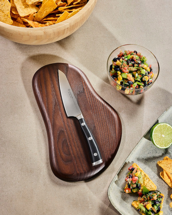 A dark wood cutting board with a uniquely shaped organic silhouette, shown with a asian utility knife resting on it. The cutting board is styled in a kitchen setting on a beige surface, surrounded by a bowl of tortilla chips, a glass bowl of salsa, and a plate with nachos and lime.