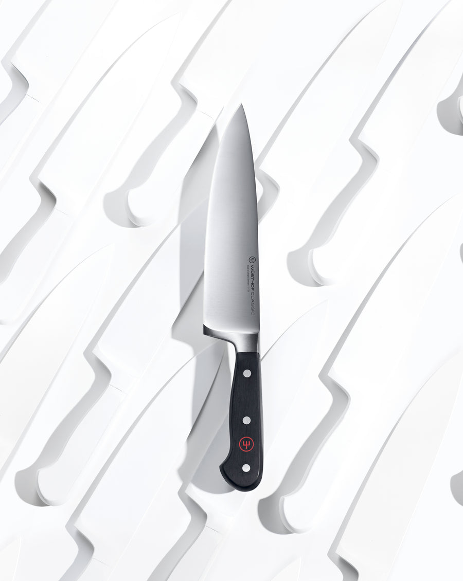 Single German chef’s knife with a black handle resting in a white molded surface shaped to hold multiple knives seen from above with high contrast lighting.