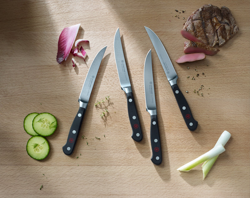 Classic 4-Piece Steak Knife Set with forged German steel blades and black triple-riveted handles arranged on a wooden cutting board with fresh ingredients.