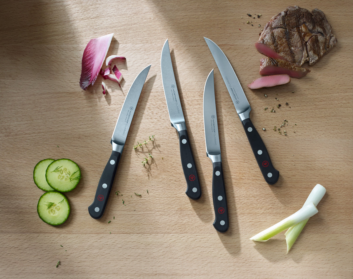Four Classic 4 1/2" Steak Knives arranged on a wooden surface with black triple-riveted handles and sharp polished stainless steel blades, accompanied by fresh vegetables and bread.
