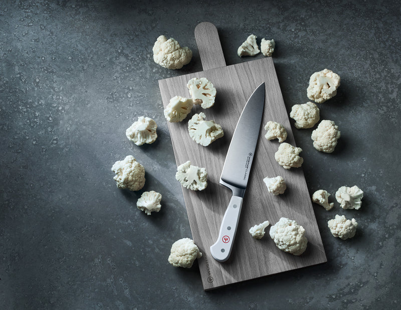 Classic 8" Chef's Knife with a polished German forged blade and white handle resting on a wooden board surrounded by cauliflower florets.