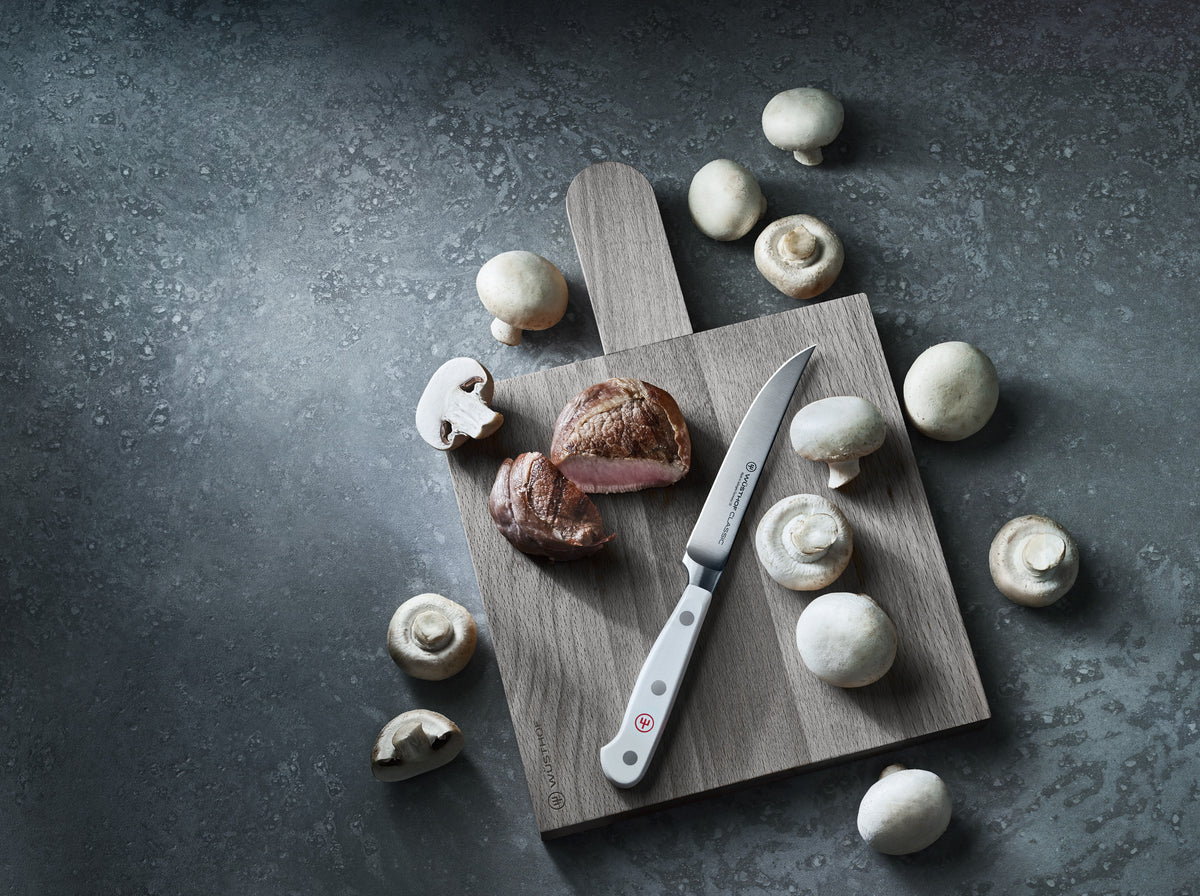Classic 4-Piece Steak Knife Set with white ergonomic handle featuring full tang and triple rivets, displayed on a wooden cutting board with sliced steak and whole mushrooms.