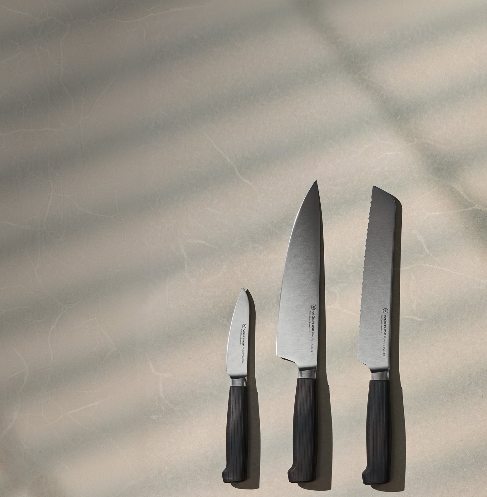 Three German kitchen knives with dark handles and stainless steel blades arranged vertically on a textured neutral surface with soft natural light and shadows.