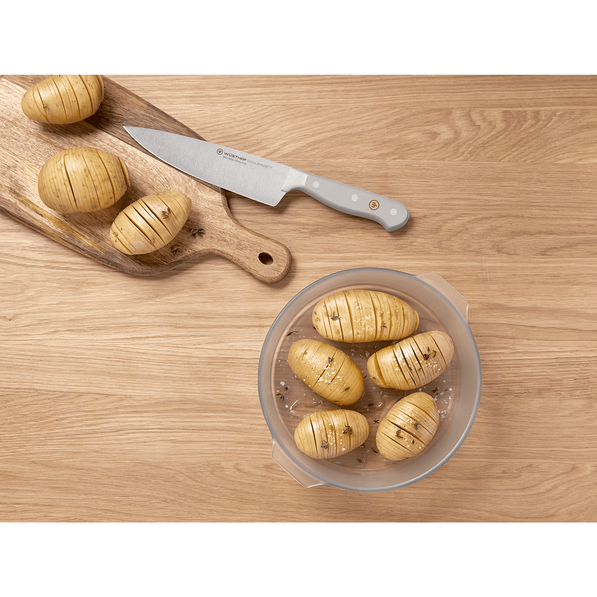 Top view of a forged German chef's knife with a grey handle from the Gourmet Grey 3-Piece Essentials Set alongside scored potatoes on a wooden surface.