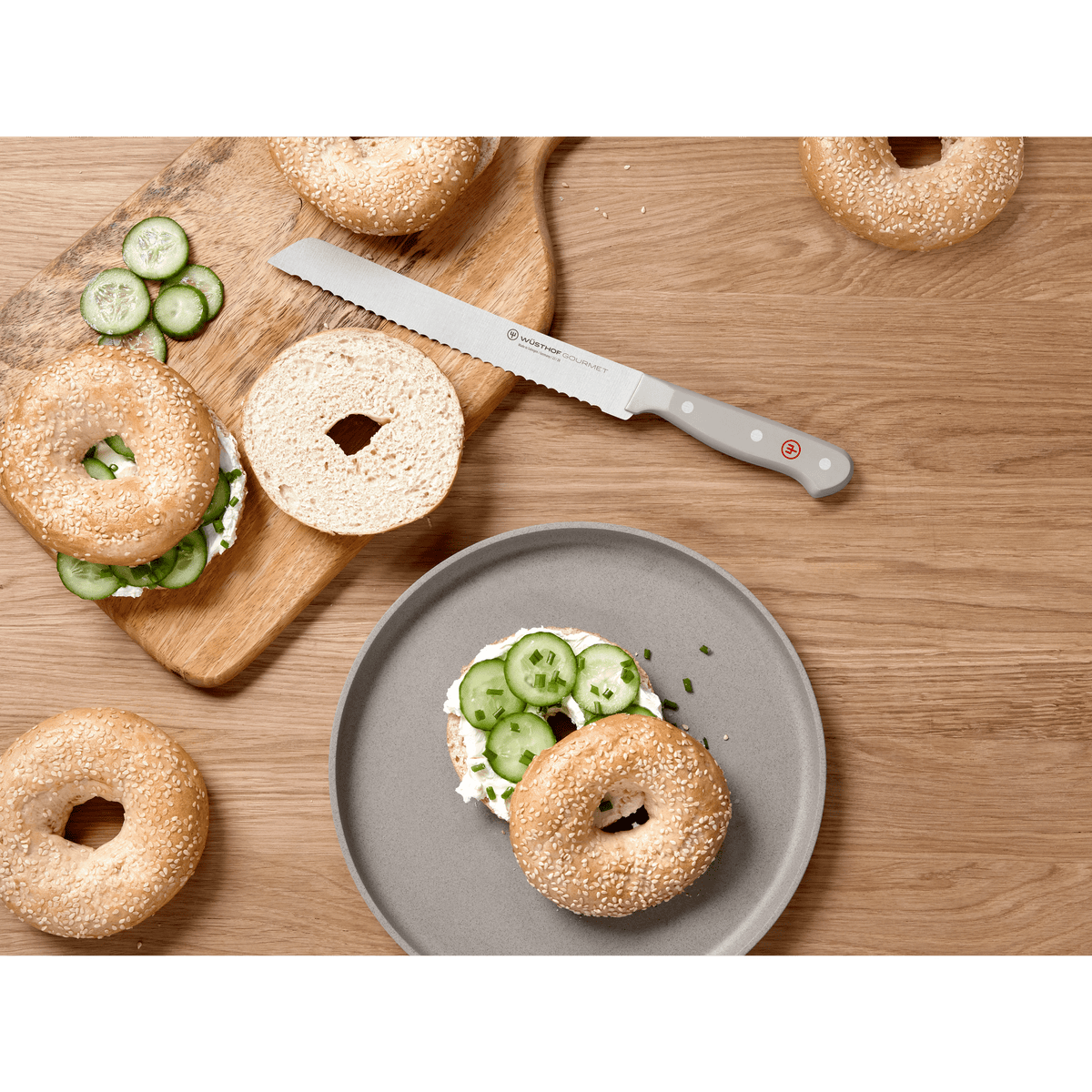 Serrated forged German bread knife with a light grey handle from the Gourmet Grey 3-Piece Starter Set placed beside sliced bagels with cream cheese and cucumber on a wooden surface.