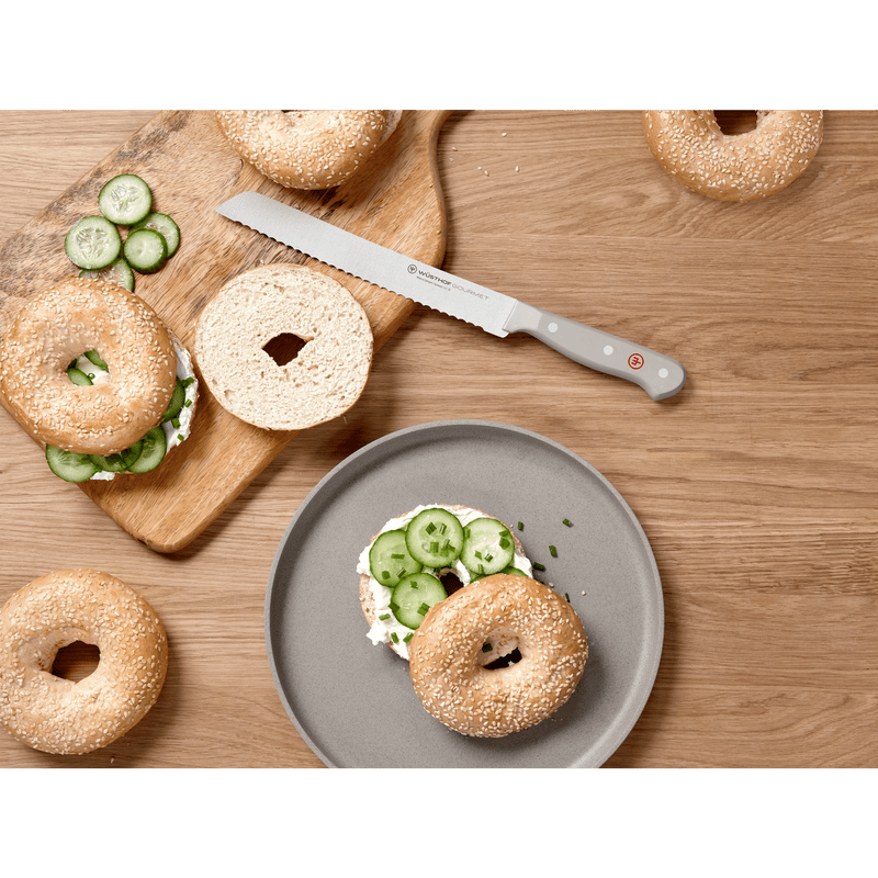 Serrated forged German bread knife with a light grey handle from the Gourmet Grey 3-Piece Starter Set placed beside sliced bagels with cream cheese and cucumber on a wooden surface.