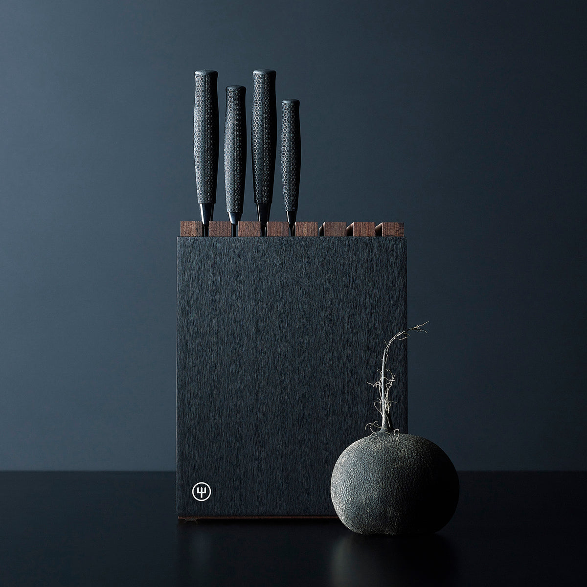 7-Slot Black Aluminum Knife Block with Wood Insert holding four knives with textured ergonomic black handles, shown with a dark textured vegetable nearby.
