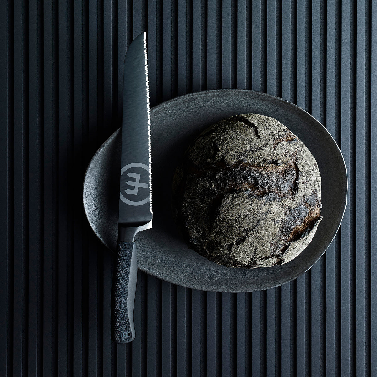 Performer 9" Precision Double-Serrated Bread Knife with matte black blade and textured ergonomic handle beside a rustic round loaf on a dark plate.