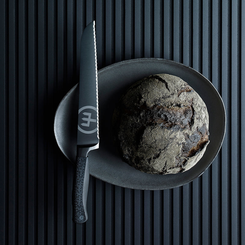 Performer 9" Precision Double-Serrated Bread Knife with matte black blade and textured ergonomic handle beside a rustic round loaf on a dark plate.