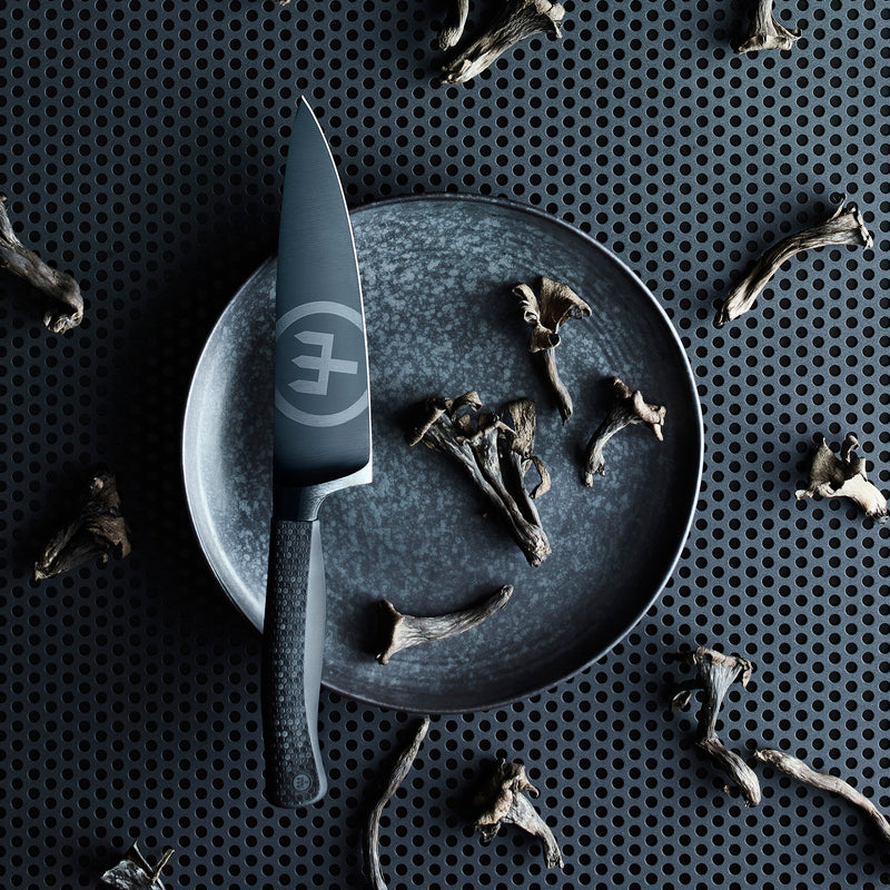 Performer 6" Chef's Knife with a black matte forged German steel blade and textured ergonomic handle displayed on a dark plate with mushrooms.