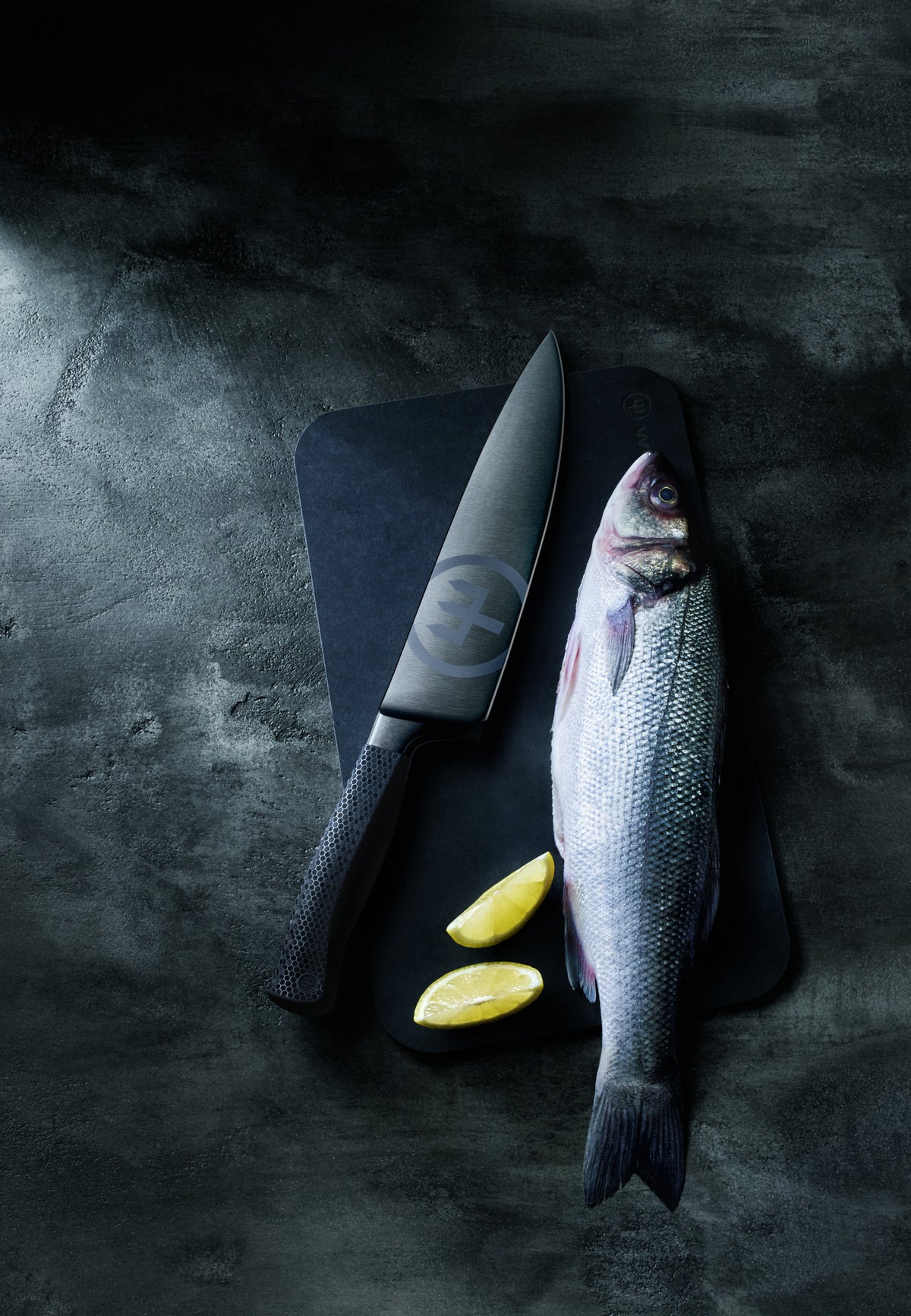 Performer 8" Chef's Knife with black forged German steel blade resting on a dark cutting board alongside a whole fresh fish and lemon wedges.