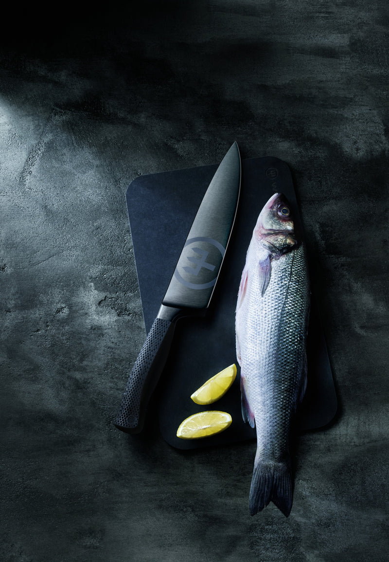 Performer 8" Chef's Knife with black forged German steel blade resting on a dark cutting board alongside a whole fresh fish and lemon wedges.