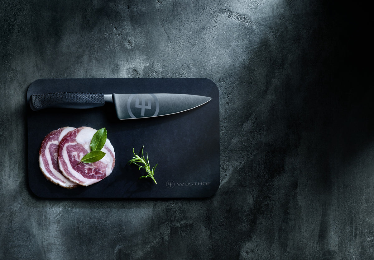Performer 6" Chef's Knife displayed on a dark cutting board with fresh herb sprigs and sliced cured meat, highlighting its forged blade and ergonomic handle.