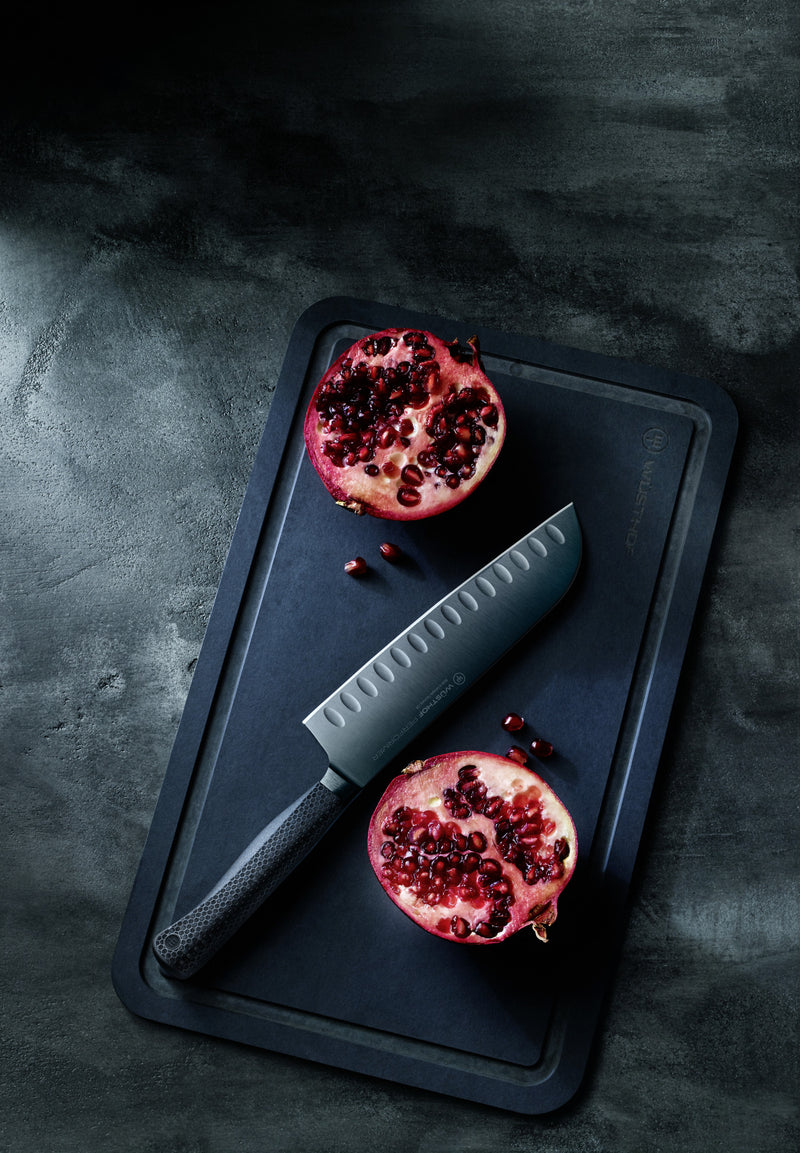 Performer 7" Santoku with matte black forged German steel blade featuring hollow edge indentations and textured handle on a dark cutting board with halved pomegranate.