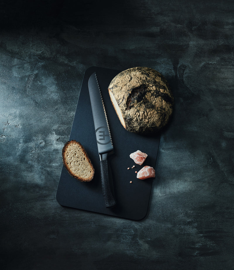 Performer 9" Precision Double-Serrated Bread Knife with a matte black blade and textured ergonomic handle displayed alongside sliced artisanal bread and coarse salt on a dark cutting board.