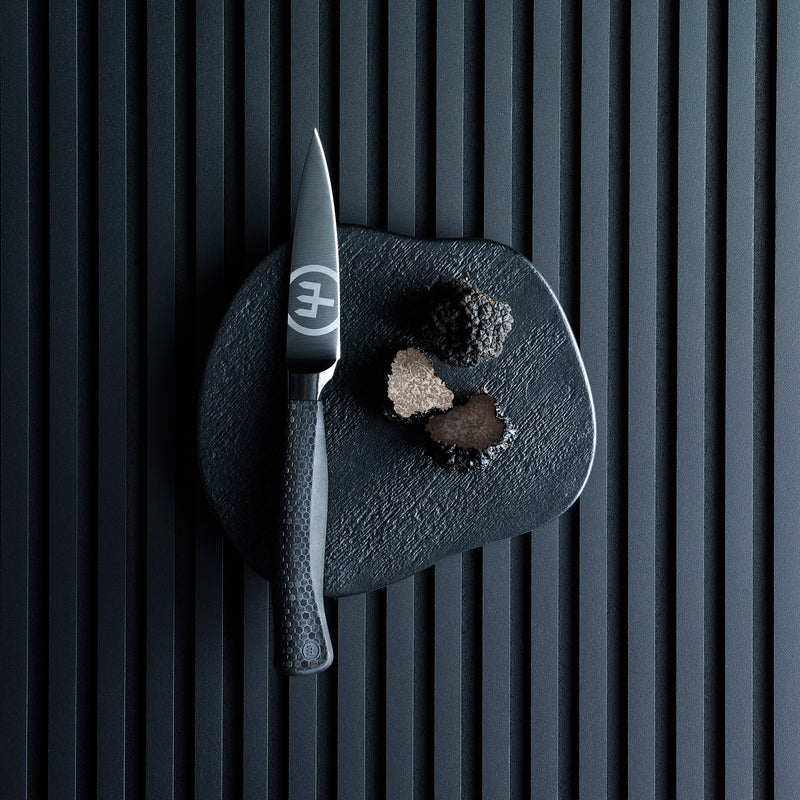 Performer 3 1/2" Paring Knife with a matte black forged blade and textured ergonomic handle displayed on a black textured board with three truffle pieces.