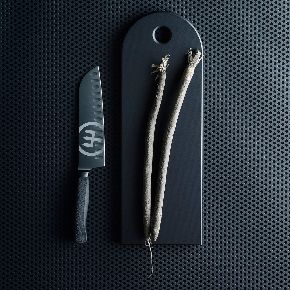 Performer 7" Santoku with forged black German steel blade and textured ergonomic handle placed beside a dark cutting board holding two root vegetables.