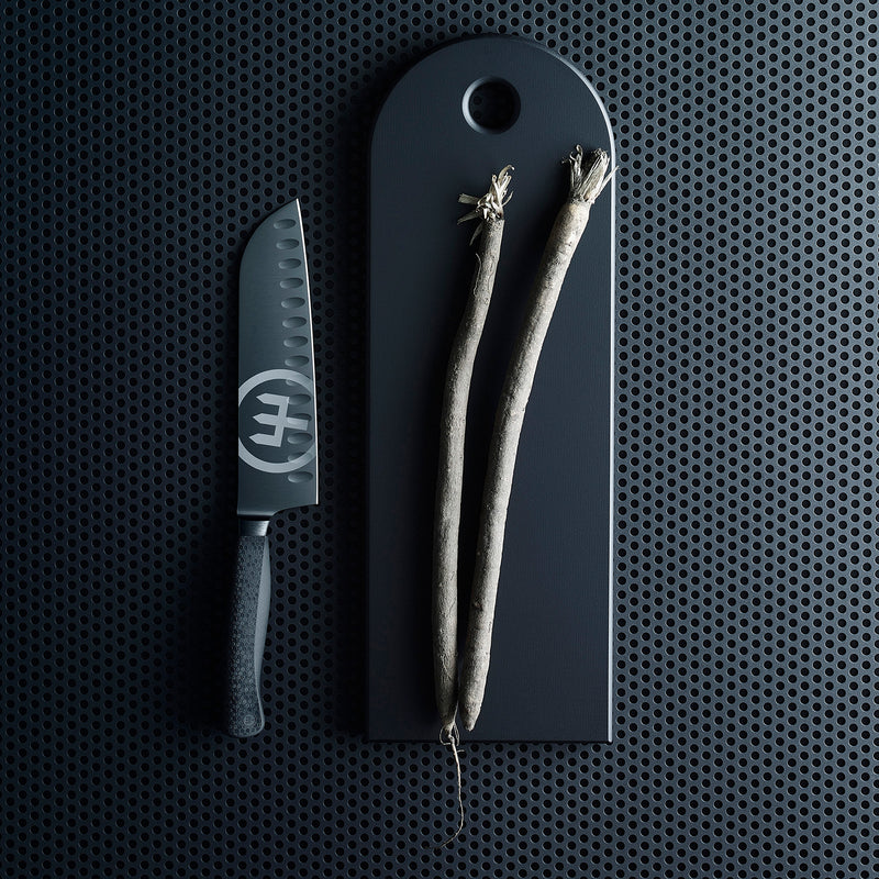 Performer 7" Santoku with forged black German steel blade and textured ergonomic handle placed beside a dark cutting board holding two root vegetables.