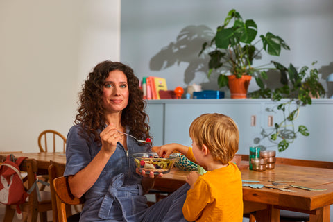 Woman with curly hair sitting at a wooden table, eating a bowl of fruit salad while a young child reaches for food nearby in a cozy dining room.