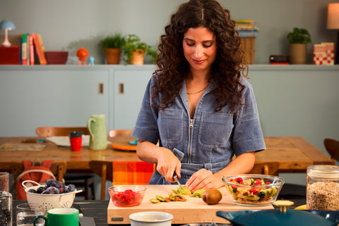 Woman with curly hair wearing a blue denim jumpsuit slicing kiwi on a wooden cutting board in a bright kitchen with bowls of fresh fruit and snacks nearby.