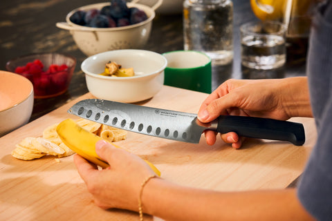 Person holding a peeled banana and preparing to slice it on a wooden cutting board using a chef’s knife with a dark handle, with bowls of fruit and glasses of water nearby.