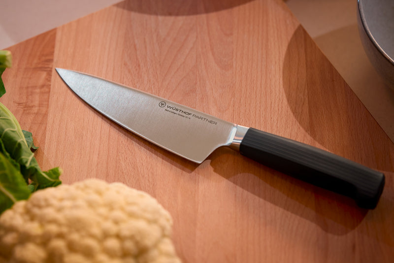 Partner 7-Piece Knife Block Set featuring a forged German chef’s knife with a textured black handle resting on a wooden cutting board next to fresh cauliflower and lettuce.
