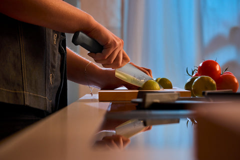Person using a forged kitchen knife with a black handle to slice green limes on a wooden cutting board, with tomatoes and more limes nearby on a kitchen counter.