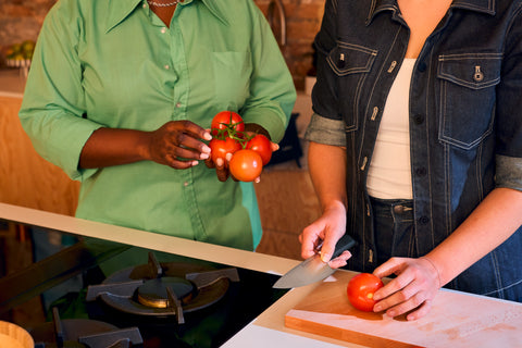 Two people preparing tomatoes in a kitchen, one holding a cluster of tomatoes and the other slicing a tomato on a wooden cutting board with a chef’s knife.