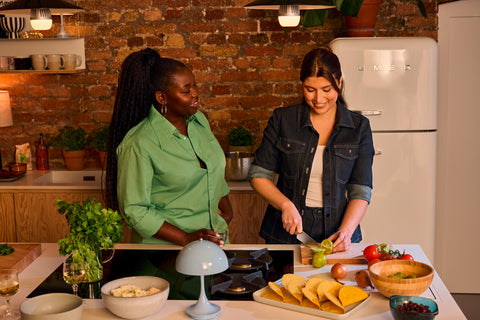 Two women chatting in a cozy kitchen, one slicing a lime with a chef’s knife while the other watches, surrounded by fresh ingredients and kitchenware.