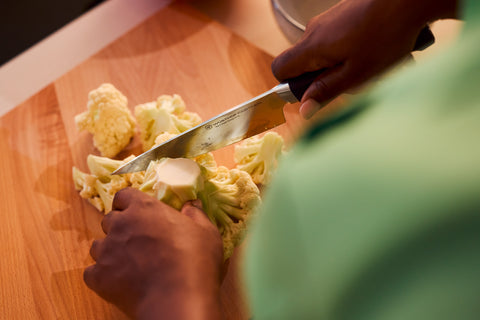 Person slicing cauliflower into florets on a wooden cutting board using a forged kitchen knife with a black handle in a home kitchen setting.
