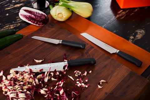 Three kitchen knives with black handles arranged on and near a wooden cutting board with chopped radicchio, fennel, and zucchini on a dark countertop.