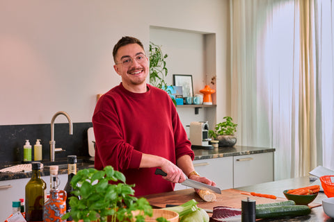 Man with glasses and a mustache wearing a red sweater slicing a baguette on a wooden board in a bright modern kitchen with plants and cooking ingredients around.