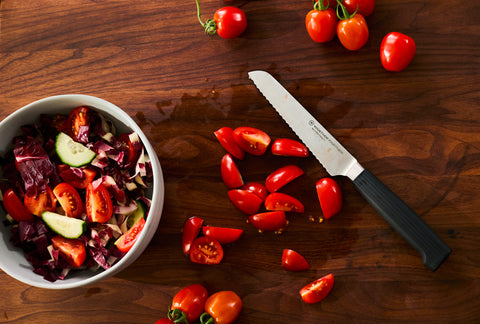 Serrated kitchen knife with a black handle on a wooden board surrounded by halved cherry tomatoes next to a bowl of mixed salad with radicchio and cucumber.