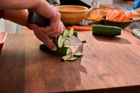 Person using a forged kitchen knife with a black handle to slice fresh cucumber on a wooden cutting board in a home kitchen setting.