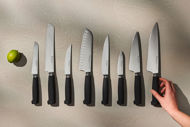 Partner 7-Piece Knife Block Set displaying nine forged German knives with black ergonomic handles arranged on a countertop, with a lime nearby and a hand reaching for the largest chef’s knife.