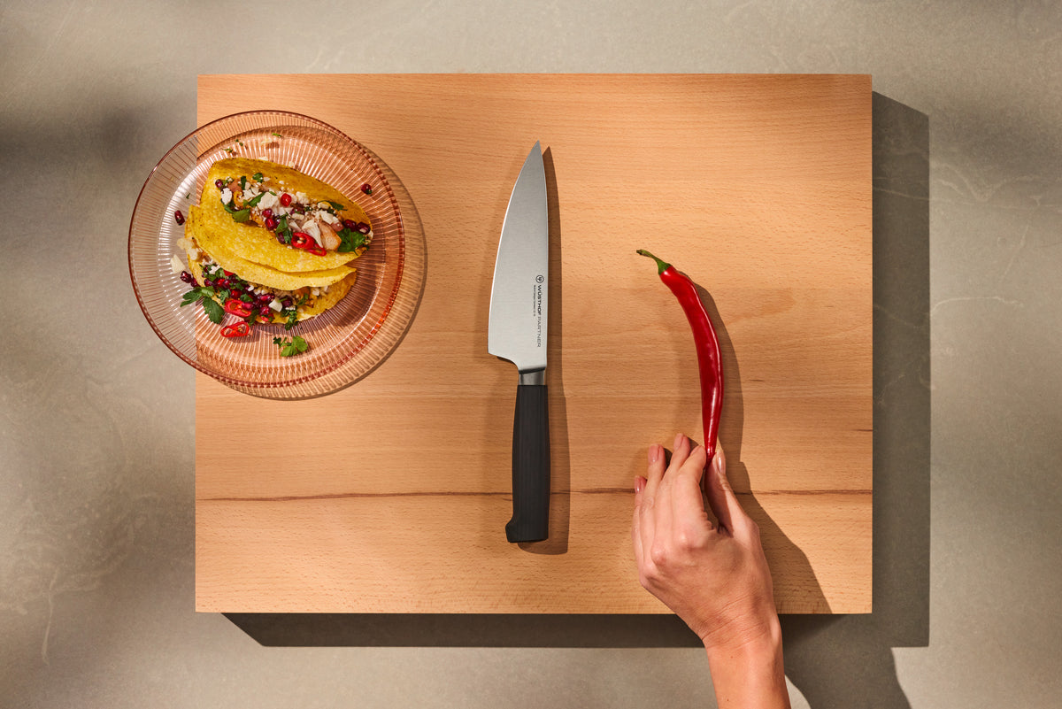 Partner 7-Piece Knife Block Set showcasing a forged German chef’s knife with a smooth black handle on a wooden cutting board alongside a fresh red chili and prepared tacos.