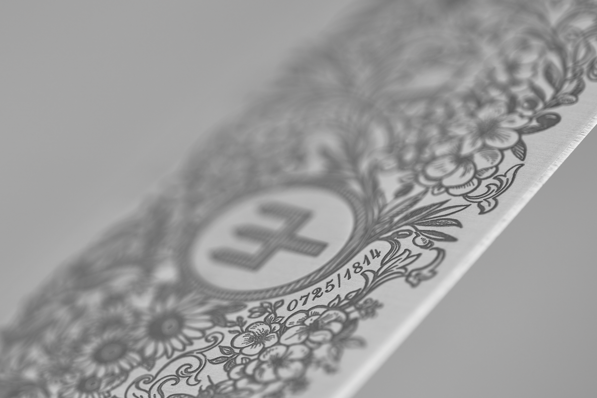 Close-up of the Amici 1814 forged kitchen knife blade featuring intricate floral engravings surrounding a central emblem and fine serial numbering.