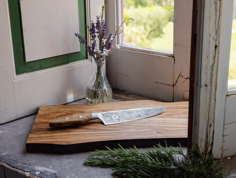 Amici 1814 forged kitchen knife with finely detailed floral blade engraving and a smooth olive wood handle resting on a wooden cutting board near a window.