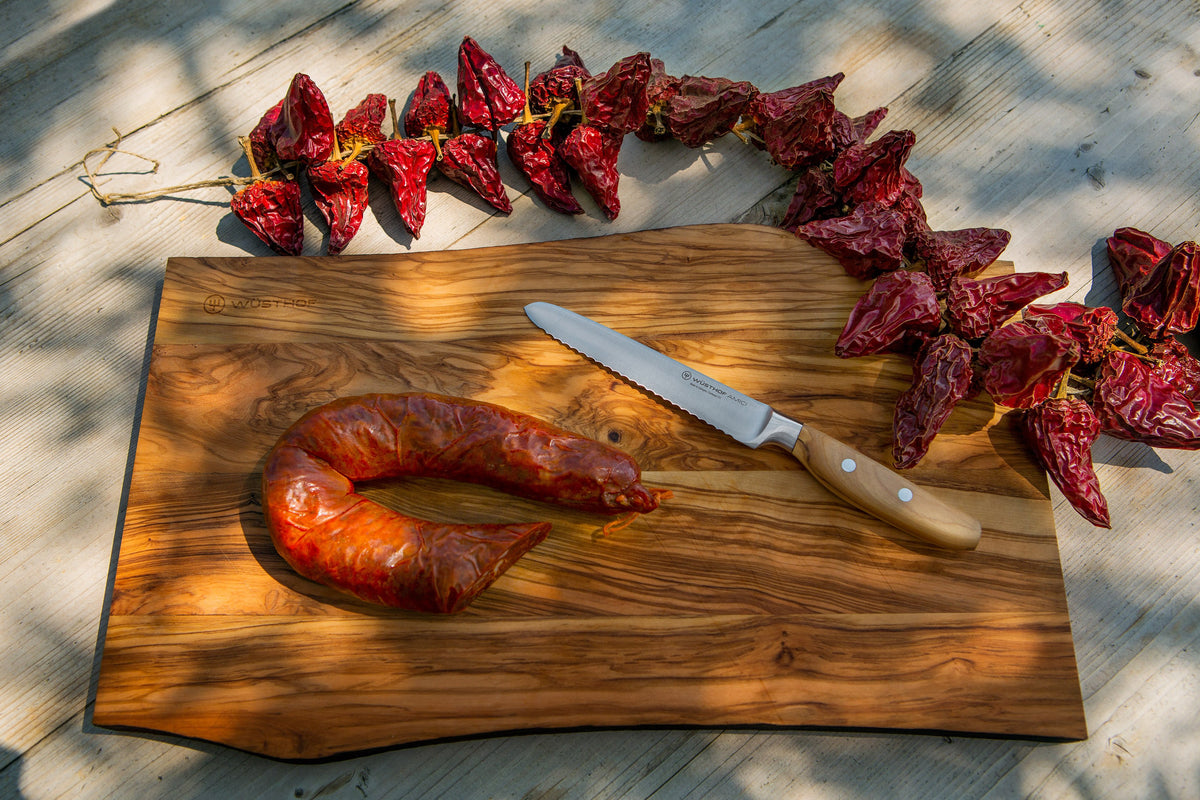 Amici 5" Serrated Utility Knife with a finely serrated blade and smooth wooden handle resting on an olive wood cutting board with smoked sausage and dried red chili peppers.