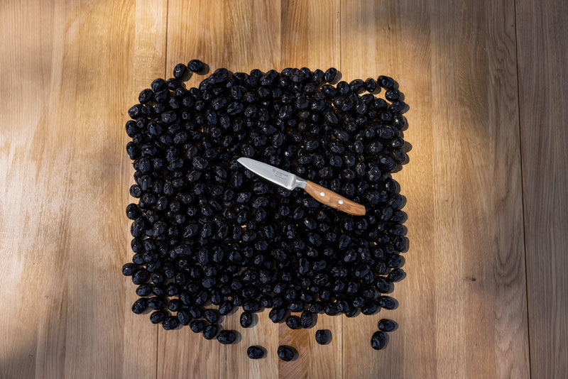 Amici 3 1/2" Paring Knife with a smooth wooden handle resting on a pile of dark dried olives on a wooden surface.