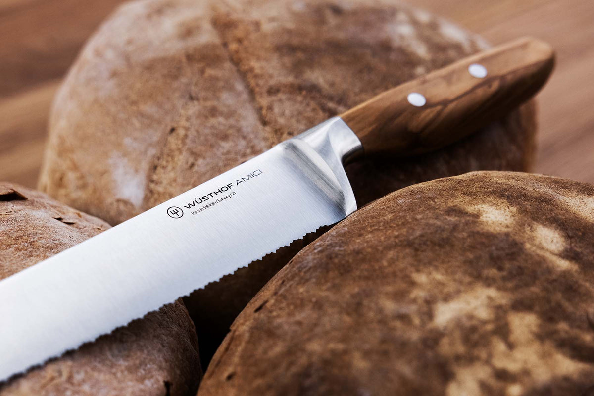 WÜSTHOF 'Amici 9' Precision Double-Serrated Bread Knife with a forged stainless steel blade and ergonomic olive wood handle resting against artisan bread loaves.