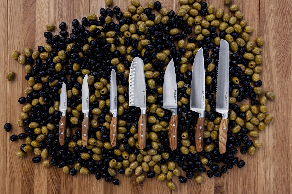 Amici 5" Serrated Utility Knife displayed alongside a collection of kitchen knives with wooden handles, arranged on a wooden surface covered with black and green olives.