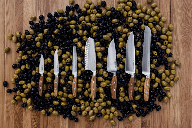 Amici 5" Serrated Utility Knife displayed alongside a collection of kitchen knives with wooden handles, arranged on a wooden surface covered with black and green olives.
