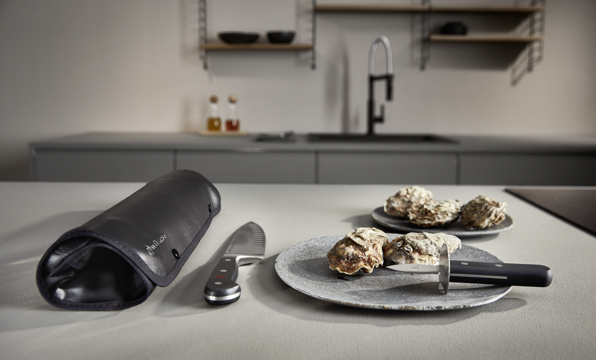 Oyster Opener with a sturdy stainless steel blade and a black ergonomic handle, displayed with oysters and a leather storage pouch on a kitchen countertop.