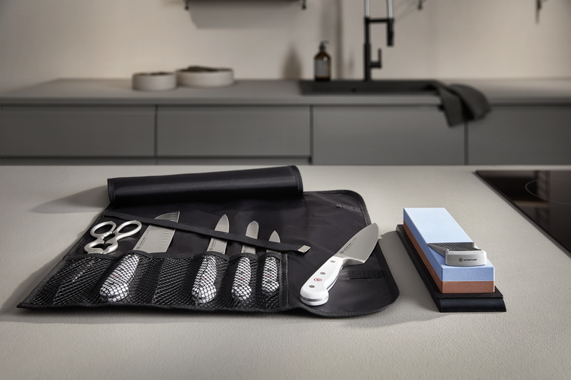 Nylon Knife Roll opened on a kitchen countertop displaying a set of forged kitchen knives, scissors, and a sharpening steel alongside sharpening stones.
